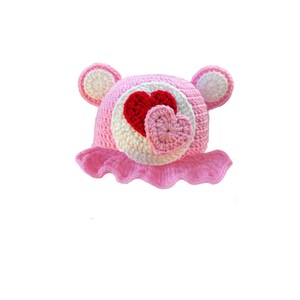 May include: Pink crocheted hat with bear ears and a heart design. The heart is made of pink and red yarn.