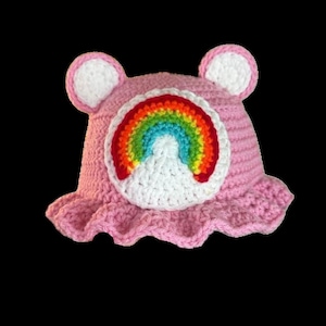May include: Pink crocheted hat with a rainbow and white cloud design. The hat has bear ears and a ruffled brim.
