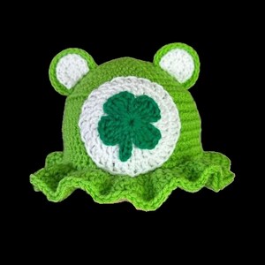 May include: Green crocheted hat with a white clover design. The hat has a ruffled brim.