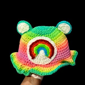 May include: A rainbow crochet bucket hat with a white crocheted rainbow design. The hat has a wide brim and is made with a variety of colorful yarns.