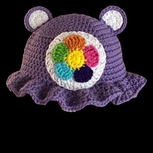 May include: Purple crochet hat with a white flower design featuring a rainbow of colors. The hat has bear ears.