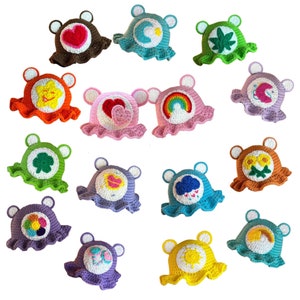 May include: A collection of 16 crocheted Care Bear hats in various colors and designs. Each hat features a different Care Bear character with their signature symbol and colors. The hats are all made with soft yarn and have a floppy brim.