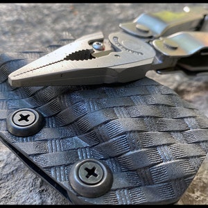 May include: A black multi-tool holster with a woven pattern and two silver screws. A silver multi-tool with pliers is partially visible in the holster.