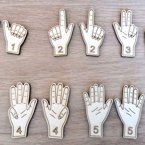 Può includere: Ritagli di legno a forma di mano, ognuno dei quali mostra un numero diverso da uno a cinque usando gesti delle dita. I ritagli sono in legno chiaro con contorni e numeri neri, ideali per scopi educativi o giochi di conteggio.