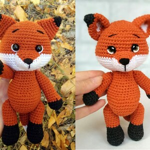 May include: A crocheted fox plush toy with orange fur, black ears, black feet, and white facial features. The toy is standing on a surface with a blurred background.