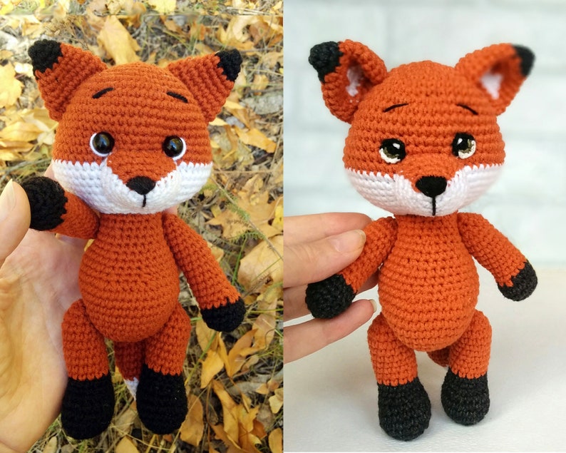 May include: A crocheted fox plush toy with orange fur, black ears, black feet, and white facial features. The toy is standing on a surface with a blurred background.