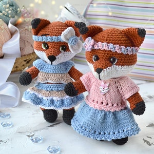 May include: Two crocheted fox plushies wearing dresses. One fox is wearing a blue and brown dress with a white bow. The other fox is wearing a pink and blue dress with a pink flower headband.