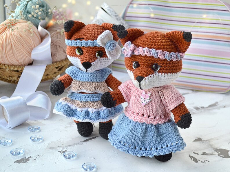 May include: Two crocheted fox plushies wearing dresses. One fox is wearing a blue and brown dress with a white bow. The other fox is wearing a pink and blue dress with a pink flower headband.
