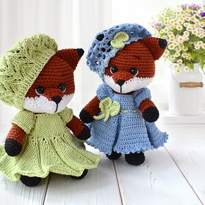 May include: Two crocheted fox dolls wearing dresses and hats. The fox on the left is wearing a green dress and a green beret. The fox on the right is wearing a blue dress and a blue hat with a white flower.