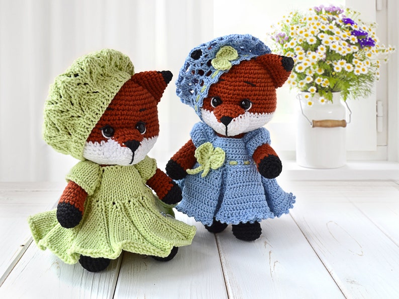 May include: Two crocheted fox dolls wearing dresses and hats. The fox on the left is wearing a green dress and a green beret. The fox on the right is wearing a blue dress and a blue hat with a white flower.