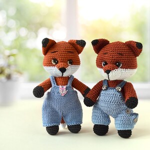 May include: Two crocheted fox plushies wearing blue dungarees. The foxes have orange fur, black noses, and white accents around their eyes and mouths.