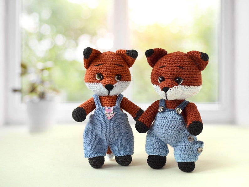 May include: Two crocheted fox plushies wearing blue dungarees. The foxes have orange fur, black noses, and white accents around their eyes and mouths.