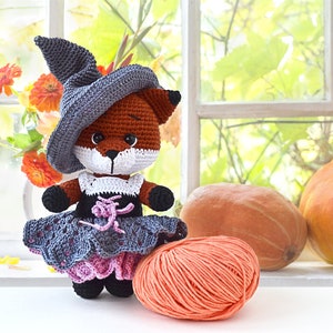 May include: A crocheted fox wearing a witch's hat and a grey and pink dress. The fox is holding a ball of orange yarn. The fox is sitting in front of a window with pumpkins in the background.