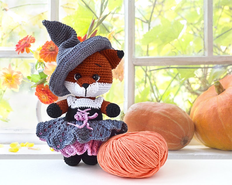 May include: A crocheted fox wearing a witch's hat and a grey and pink dress. The fox is holding a ball of orange yarn. The fox is sitting in front of a window with pumpkins in the background.