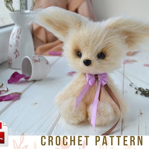 May include: Crochet pattern for a beige amigurumi fox with a pink bow. The fox is sitting on a white wooden surface.