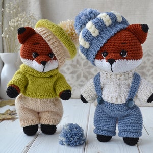 May include: Two crocheted fox plushies wearing different outfits. One fox is wearing a green sweater and beige trousers, while the other is wearing a white sweater and blue overalls. Both foxes are wearing hats.