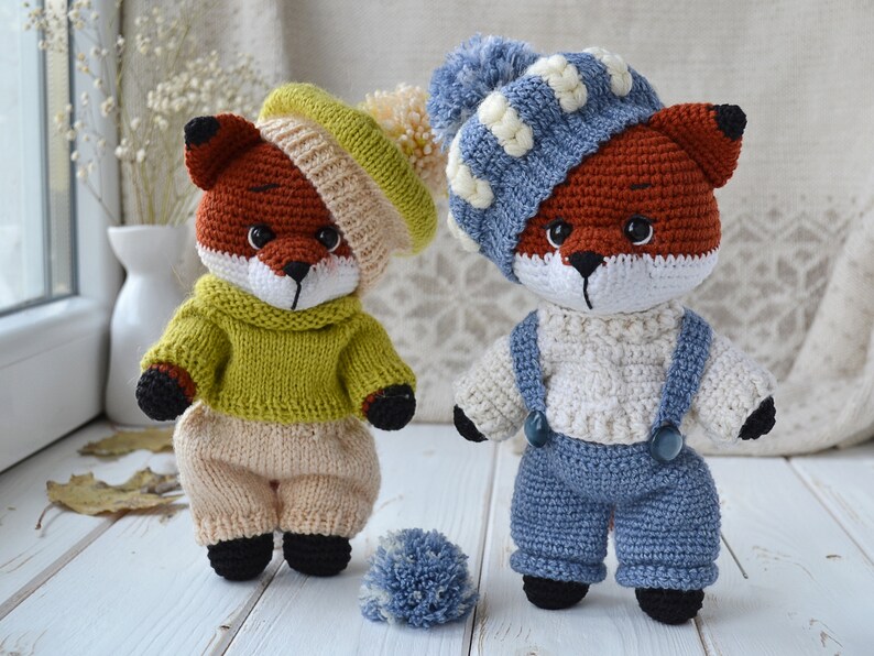 May include: Two crocheted fox plushies wearing different outfits. One fox is wearing a green sweater and beige trousers, while the other is wearing a white sweater and blue overalls. Both foxes are wearing hats.