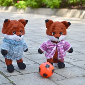May include: Two crocheted fox plushies, one wearing a blue sweater and the other wearing a pink and white striped sweater with a pink ribbon, playing with an orange football on a brick pathway.