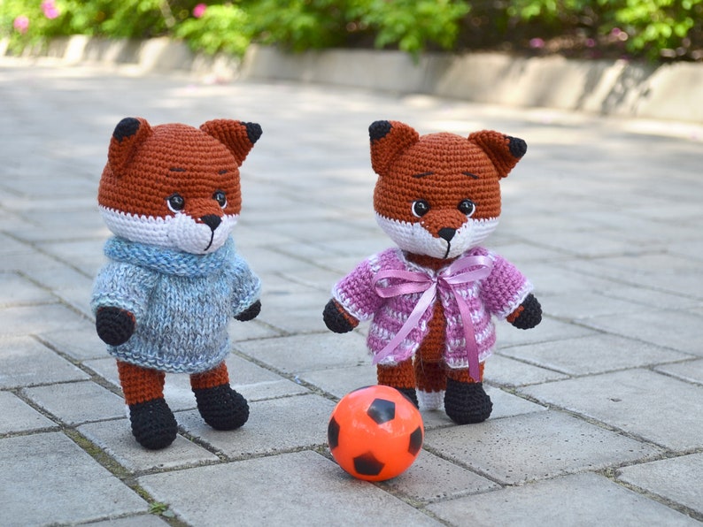 May include: Two crocheted fox plushies, one wearing a blue sweater and the other wearing a pink and white striped sweater with a pink ribbon, playing with an orange football on a brick pathway.
