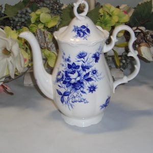 Op de afbeelding: Witte porseleinen theepot met een blauw bloemmotief. De theepot heeft een gebogen tuit, een decoratieve handgreep en een bijpassende deksel. Het bloemmotief omvat rozen en andere bloemen.