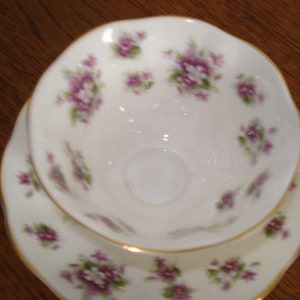 May include: A white teacup and saucer set with a delicate floral pattern in shades of purple and green. The set features a gold trim around the edges.