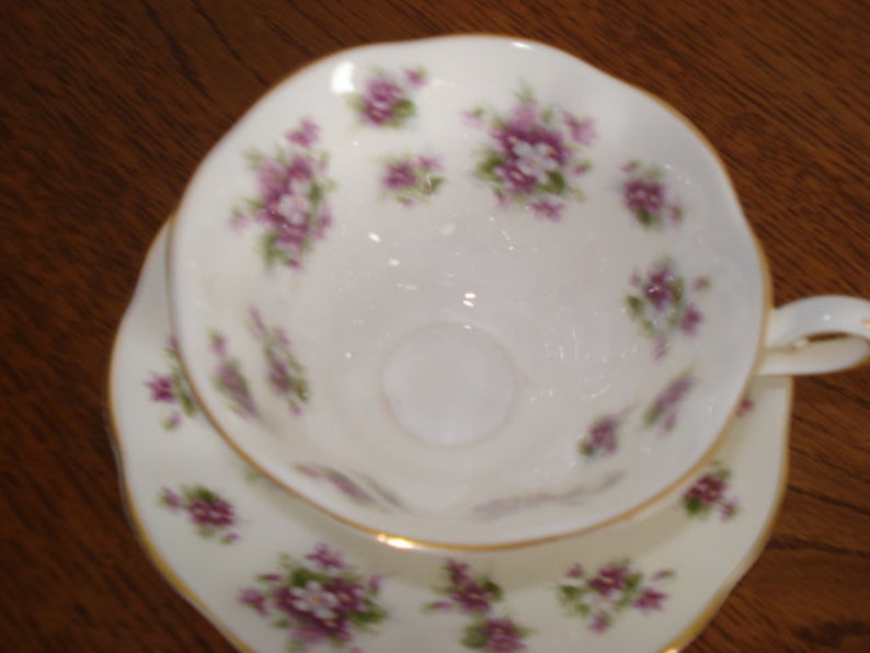 May include: A white teacup and saucer set with a delicate floral pattern in shades of purple and green. The set features a gold trim around the edges.