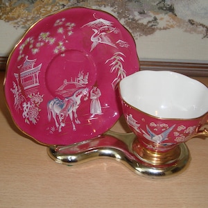 Puede incluir: Un juego de taza y platillo de té rosa con ribete dorado y un diseño floral blanco. El platillo presenta una escena de un caballo y un jinete en un paisaje.