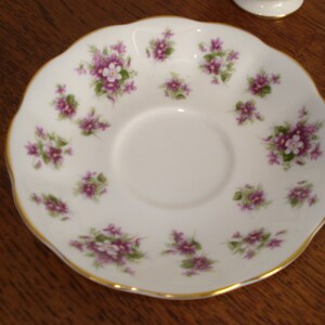 May include: A white porcelain saucer with a gold rim and a delicate floral pattern of purple and white flowers. The saucer is designed for a teacup.