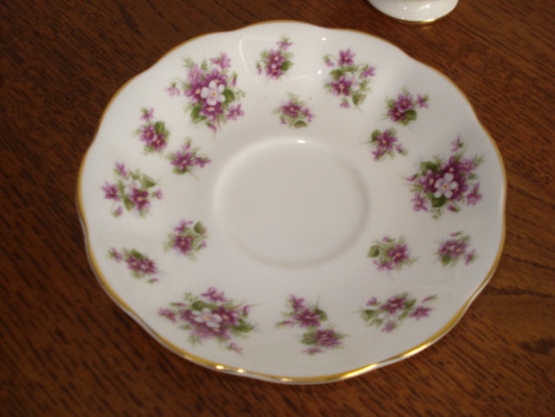 May include: A white porcelain saucer with a gold rim and a delicate floral pattern of purple and white flowers. The saucer is designed for a teacup.
