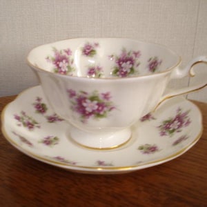 May include: A white teacup and saucer with a floral pattern of purple and green flowers. The cup has a delicate handle and a scalloped edge. The saucer has a matching floral pattern.