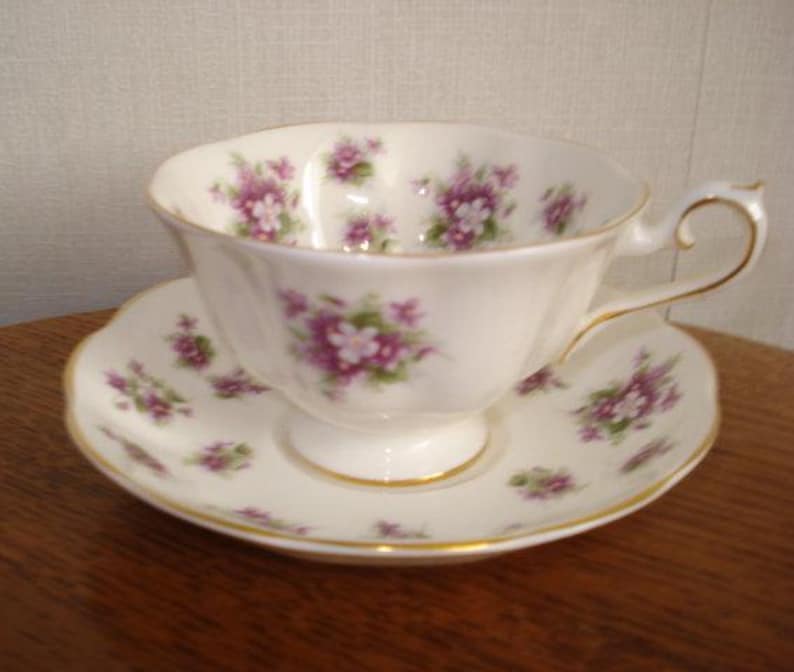 May include: A white teacup and saucer with a floral pattern of purple and green flowers. The cup has a delicate handle and a scalloped edge. The saucer has a matching floral pattern.