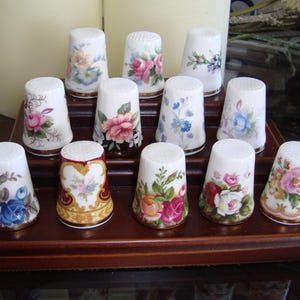 Könnte beinhalten: Ein Set mit 10 Porzellan-Fingerhüten mit Blumendesigns. Die Fingerhüte sind in einer mehrstufigen Anzeige auf einem Holzständer angeordnet. Jeder Fingerhut zeigt ein anderes Blumenmuster, darunter Rosen, Gänseblümchen und andere Wildblumen.