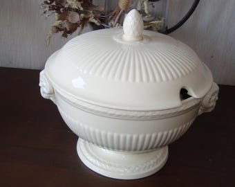 May include: A cream-colored ceramic tureen featuring a ribbed body and lid, a finial shaped like a pinecone, and two side handles with decorative scroll details. The lid includes a small cutout for a ladle, and the base is accented with a rope pattern.