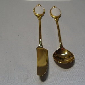 Può includere: Un coltello da burro dorato e un cucchiaino. Entrambi gli utensili hanno un design ovale bianco sulla parte superiore del manico. Il coltello ha una lama curva e il cucchiaino ha una forma arrotondata. Il set è probabilmente per servire.