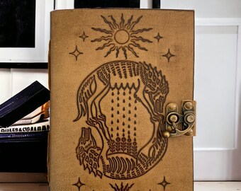 Pode incluir: Um diário de couro marrom com um design estampado de dois lobos uivando para a lua. O diário tem um fecho de latão e é decorado com estrelas e sóis.