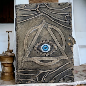 Op de afbeelding: Een bruin leren dagboek met een gouden reliëf van een driehoek en een blauw oog in het midden. Het dagboek heeft een sluiting met een gesp.