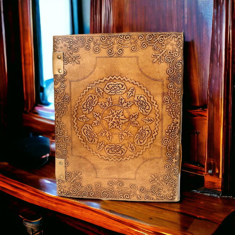 Puede incluir: Un diario de cuero marr&oacute;n con intrincados dise&ntilde;os florales en relieve en la portada. El diario tiene cierres de lat&oacute;n y un borde decorativo.