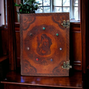 May include: A brown leather journal with a mermaid design and colorful gemstones. The journal has two brass clasps.