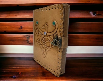 May include: Tan leather journal with a Celtic dragon design. The cover features an embossed dragon and knotwork, accented with green and blue gemstones. The journal has a stitched border and a metal clasp closure.