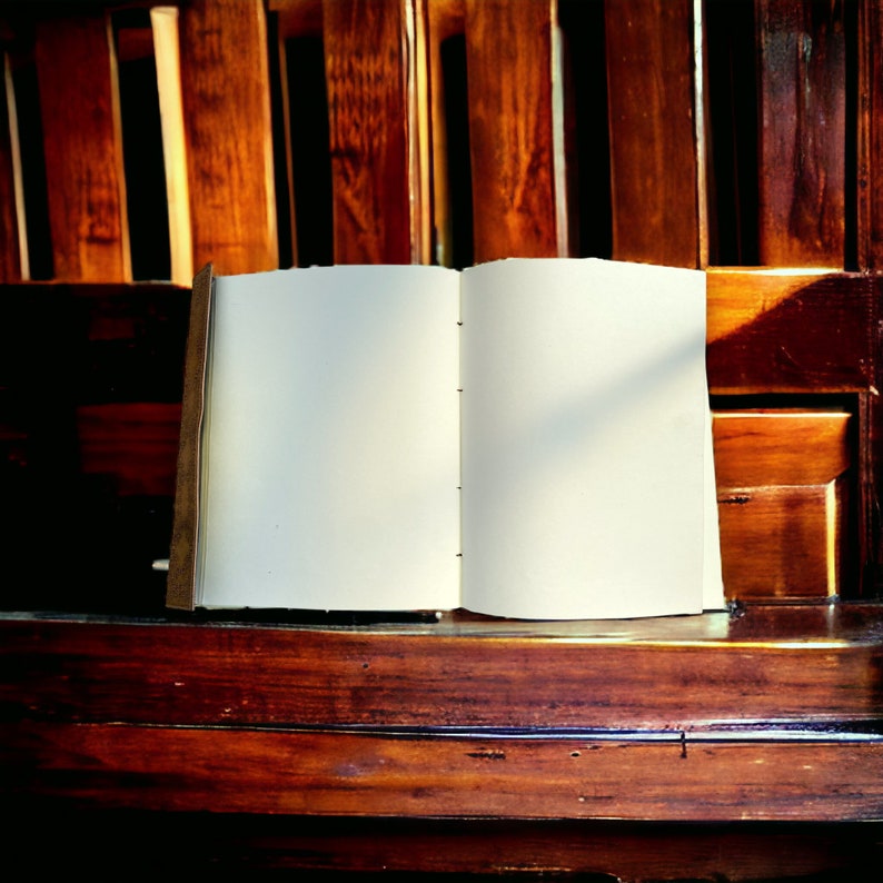 Puede incluir: Un cuaderno abierto en blanco con una cubierta de cuero marr&oacute;n se encuentra sobre una superficie de madera. El cuaderno est&aacute; abierto en una p&aacute;gina en blanco con una sombra proyectada en el lado derecho de la p&aacute;gina.