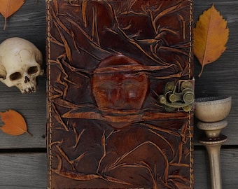 Pode incluir: Um diário de couro castanho com uma aparência texturizada e enrugada. O diário apresenta um design central em relevo e um fecho de metal. A imagem inclui uma caveira, folhas de outono e um objeto decorativo de metal, sugerindo um tema gótico ou outonal.