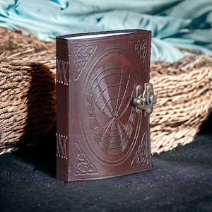May include: A brown leather journal with a Celtic knot design and a spider web embossed on the front cover. The journal has a brass clasp.