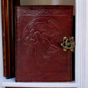 May include: A brown leather journal with a wolf howling at the moon design embossed on the front cover. The journal has a brass clasp closure.