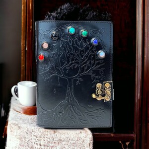 Puede incluir: Diario de cuero negro con un diseño de árbol de la vida y piedras preciosas de colores. El diario tiene un cierre de latón.