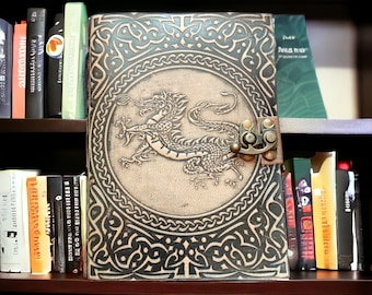 May include: A black leather journal with a Celtic knot design and a dragon embossed on the front cover. The journal has a metal clasp and is on a bookshelf.
