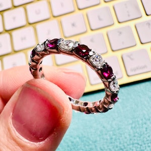 May include: A silver eternity ring featuring alternating round-cut, vibrant red gemstones and clear, sparkling white stones. The ring is held up against a blurred background of a gold-colored keyboard.