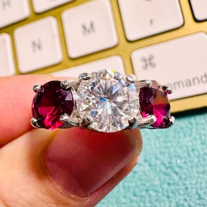 Op de afbeelding: Een zilveren ring met een grote, heldere, rond geslepen centrale steen, geflankeerd door twee kleinere, ovaal geslepen, dieprode edelstenen. De ring wordt getoond tegen een gouden toetsenbord.