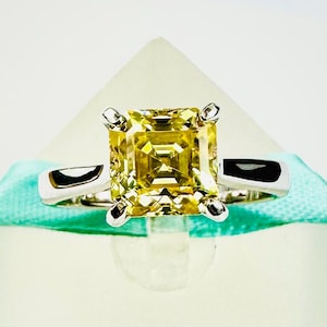 May include: A square-cut, yellow gemstone ring with a silver band. The gemstone is the focal point, set with four prongs. The ring is displayed on a light blue ribbon against a white background. The ring's design is simple yet elegant.