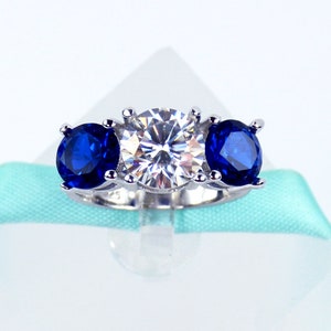 May include: A silver ring with a large, clear center stone and two smaller, blue stones on either side.