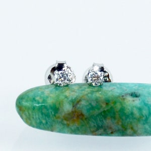 May include: A pair of silver stud earrings with a small, clear diamond in the center of each earring. The earrings are sitting on a green and brown stone.